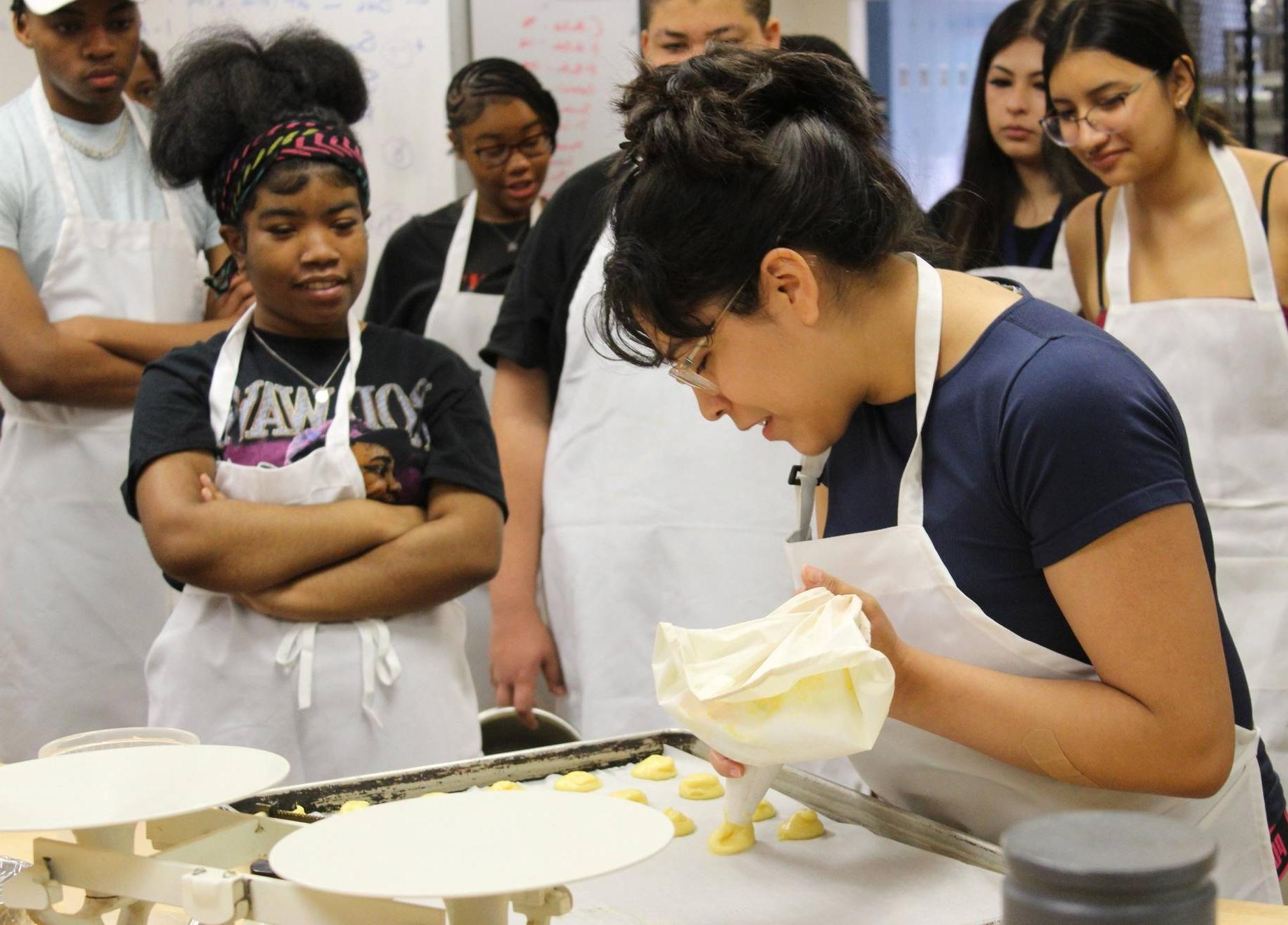 A multicultural group of teenagers wearing aprons look on as a female wearing an apron and holding a piping bag pipes dough onto a cookie sheet.